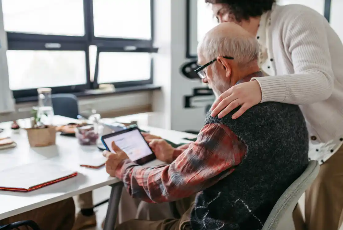Older man receiving help from a carer while looking at information on a tablet.