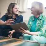 Younger woman and older lady sitting together on a sofa, smiling and talking while looking at a tablet during a care discussion