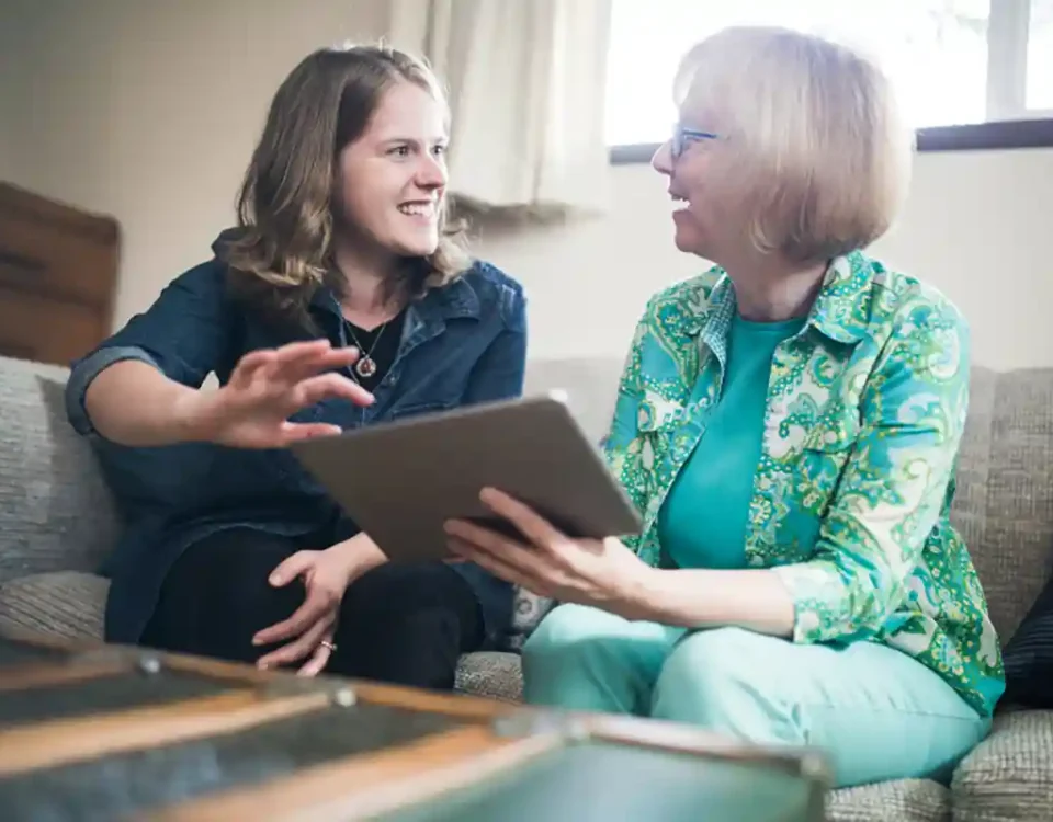 Younger woman and older lady sitting together on a sofa, smiling and talking while looking at a tablet during a care discussion
