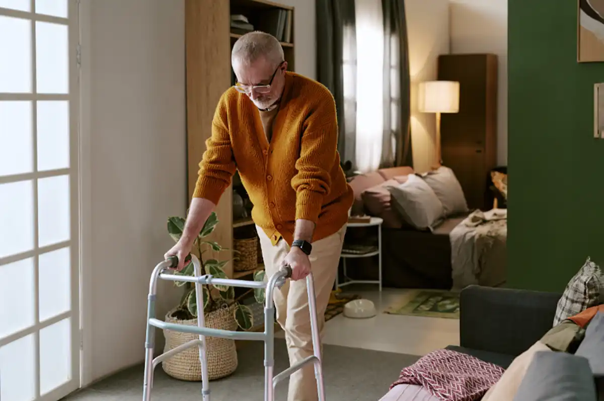 Older man using a walking frame to move around his living room while waiting for hospital treatment.