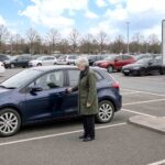 An older woman locking her car in a supermarket car park, with the parking information sign behind her.
